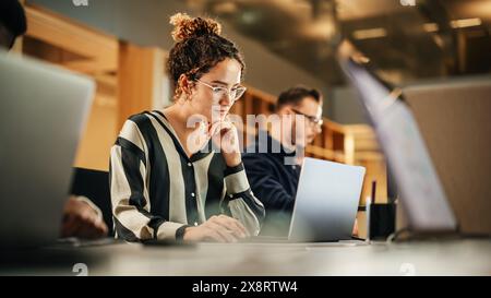 Portrait of Enthusiastic Hispanic Young Woman Working on Computer in a Modern Bright Office. Confident Human Resources Agent Smiling Happily While Collaborating Online with Colleagues. Stock Photo