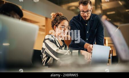 Two Colleagues Working On Laptop in Office. Female Hispanic Help Desk Coordinator Collaborates with White Male Customer Service Agent, They Discuss a Project, Chat, Smile. Teamwork Collaboration Stock Photo