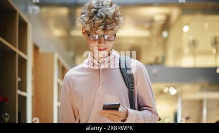 Portrait of a Young Caucasian Man Walking Across Corporate Office Hall, Browsing Web on his Smartphone and Smiling. Succesfull Entrepreneur Walks and Thinks of Startup Strategy. Stock Photo