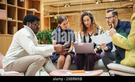 Diverse People in an Office Collectively Brainstorming and Working as a Team Using Laptop, Tablet and Charts. Group Of Multiethnic Young E-business specialists Discussing the Best Strategy Stock Photo