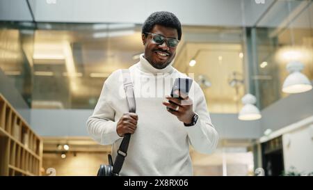Portrait of Young Black Man Feeling Confident as He Walks Smiling and Checking His Phone, in a Busy Office Hallway. He is Browsing the Internet on His Smartphone. Low Angle Stock Photo