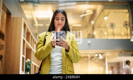 Portrait of Hispanic Businesswoman Using Smartphone App to Share Social Media Post about Career Growth. Professional Female Manager Walking Through Office Building, Smiling and Looking Happy. Stock Photo