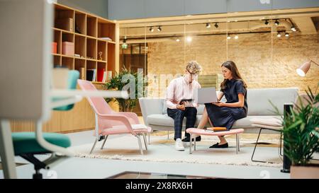 Portrait of Two Creative Colleagues Collaborating and Pointing at the Laptop in Modern Meeting Room. White Female Chief Manager Working and Advising a Young Software Engineer. Busy Office Stock Photo