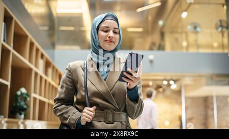 Muslim Woman wearing Hijab Checking Email App on her Smartphone After a Succesful Meeting. Portrait of a Female Content Manager Walking Through a Busy Workplace Hall. Low Angle Stock Photo