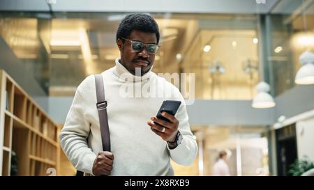 Portrait of a Black Man Walking in Corporate Office while Scrolling on his Smartphone. A Businessman Smiling While He Heads Towards His Desk in the Morning to Start a Work Day Stock Photo