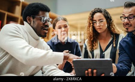 Multiethnic Young Group of People Brainstorming Together in a Meeting Room at the Office. Black Male E-commerce Coordinator Pitching His Ideas To Startup Management Board Members Using Tablet Stock Photo