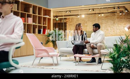 Portrait of Two Creative Colleagues Collaborating and Pointing at the Laptop Screen in Office Hub. Hispanic Young Woman Discussing Work with Black Male Assistant Stock Photo
