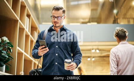 Portrait of White Man Walking in Office Hall With Smartphone and Coffee and Smiling. Male Software Developer Reading Feedback on His Work with Confidence. Low Angle, Close Up Shot Stock Photo