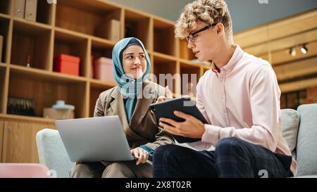 Two Creative Colleagues Discussing and Pointing at a Laptop in Modern Casual Office. Caucasian Male Sales Manager Discussing Project Plan on Tablet with Muslim Female Legal Assistant. Stock Photo