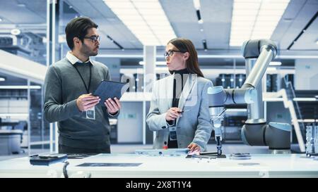 Two Industrial Robotics Engineers Gathered at a Table With Robotic Arm. Female and Male Scientists Use Tablet Computer to Manipulate and Program the Robot to Pick Up and Move a Metal Component. Stock Photo