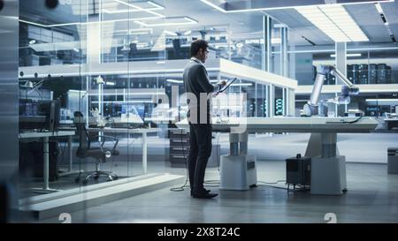 Young Multiethnic Male Engineer Using a Tablet Computer, Researching and Developing a Futuristic Robotic Arm Machine in a High Tech Industrial Laboratory with Modern Equipment. Stock Photo