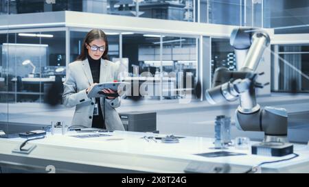 Young Beautiful Female Engineer Using a Tablet Computer, Researching and Developing a Futuristic Robotic Arm Machine in a High Tech Industrial Laboratory with Modern Equipment. Cinematic Footage. Stock Photo