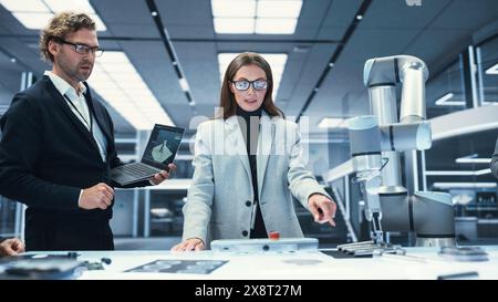 Team Leader Working with Robotics Engineers on a Robot Arm Project, Discussing the Project in Scientific Technology Lab. Young Team Researching and Developing an AI Robotic Arm in Industrial Facility. Stock Photo