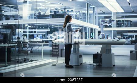 Young Female Engineer Using a Tablet Computer, Researching and Developing a Futuristic Robotic Arm Machine in a High Tech Industrial Laboratory with Modern Equipment. Stock Photo
