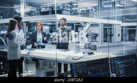 Diverse Team of Industrial Robotics Engineers Gathered Around a Table With Robotic Arm. Scientists Use Tablet Computer to Manipulate and Program the Robot to Pick Up and Move a Metal Component. Stock Photo
