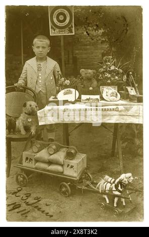 Original early 1900's postcard of young boy, wearing a smart sailor suit, displaying his toys outside in his garden, Birthday presents perhaps. Toys include teddy, soft toy dog, horse cart,  boy is Paul F maybe. Dated 23 June 1911 on front of postcard. Posted to London from Germany. Victorian boy boys /  Edwardian boy boys. Victorian child. Stock Photo
