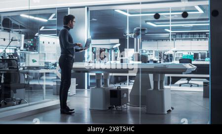 Industrial Robotics Engineer Standing Next to Robotic Arm in a High Tech Factory. Male Scientist Uses Laptop Computer to Manipulate and Program the Robot to Pick Up and Move a Microchip. Stock Photo