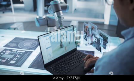 Over the Shoulder Shot of a Robotics Engineer Using a Laptop Computer with a Software Operating the Robotic Arm. Robot Hand Holding a Microchip. Automation Startup Research and Development Office. Stock Photo