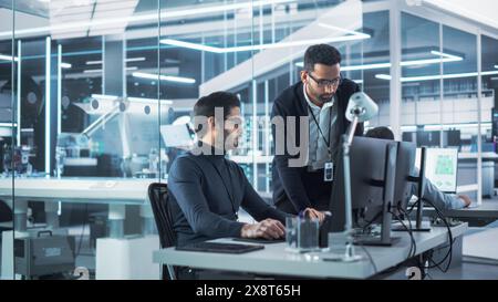 Two Industrial Engineers Discussing Work Projects, Talk and Use Desktop Computer in Robotics Startup Facility. Specialists and Professionals Researching and Developing High Tech Robotics Equipment. Stock Photo