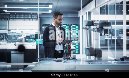 Industrial Robotics Professional Interacting with Robotic Arm During a Research Phase in a High Tech Startup. Scientist Uses Tablet Computer to Manipulate and Program the Robot to Move a Microchip. Stock Photo