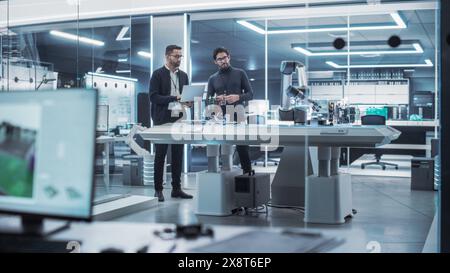 Young Engineer Works with Colleague Robotics Scientist with Laptop Computer. They Collaborate on a Robot Arm Development in an Industrial Startup. Analyzing and Evaluating Technology Systems. Stock Photo