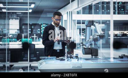 Industrial Robotics Professional Interacting with Robotic Arm During a Research Phase in a High Tech Startup. Scientist Uses Tablet Computer to Manipulate and Program the Robot to Move a Microchip. Stock Photo