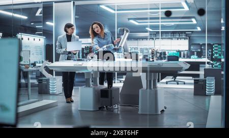 Two Female Working in High Tech Facility. Robotics Engineer and Machinery Operator Collaborate on a Robot Arm Project, Using a Laptop Computer to Operate the Robot Arm in Technology Startup Lab. Stock Photo