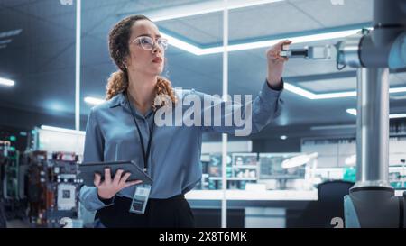 Portrait of a Young Multiethnic Female Engineer Using Tablet Computer. Interacting with a Robotic Arm Machine, Receiving a Microchip from the Hand. High Tech Industrial Laboratory Facility. Stock Photo