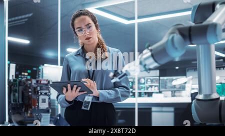 Industrial Robotics Engineer Interacting with Robotic Arm During an Experiment in a High Tech Startup. Female Scientist Uses Tablet Computer to Manipulate and Program the Robot to Move a Microchip. Stock Photo