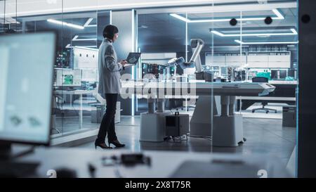 Industrial Robotics Engineer Standing Next to a Robotic Arm in a High Tech Factory. Female Scientist Uses Laptop Computer to Manipulate and Program the Robot to Pick Up and Move a Microchip. Stock Photo