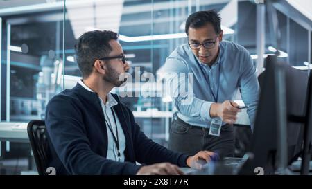 Two Industrial Engineers Discussing Work Projects, Talk and Use Desktop Computer in Robotics Startup Facility. Specialists and Professionals Researching and Developing High Tech Robotics Equipment. Stock Photo