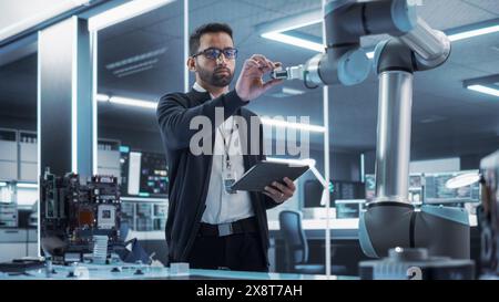 Portrait of a Young Multiethnic Male Engineer Using Tablet Computer. Interacting with a Robotic Arm Machine, Receiving a Microchip from the Hand. High Tech Industrial Laboratory Facility. Stock Photo
