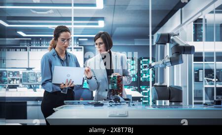 Two Female Working in High Tech Facility. Robotics Engineer and Machinery Operator Collaborate on a Robot Arm Project, Using a Laptop Computer to Operate the Robot Arm in Technology Startup Lab. Stock Photo
