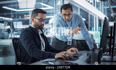 Male Project Supervisor Talks to an Industry 4 Engineer Who Works on Desktop Computer in Robotics Startup Industrial Office. Software Developer Discussing Work Related Tasks During a Meeting. Stock Photo