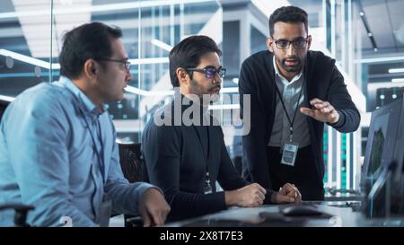 Team of Three Handsome Talented Young Engineers Use Computer to Discuss a Technological Project in Industry 4.0 Modern Factory Office. Group of Scientists Work in Research and Development Center. Stock Photo