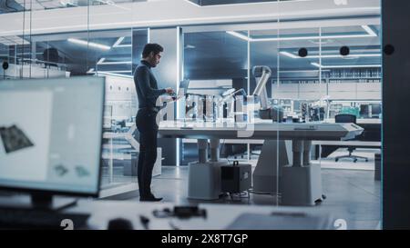Industrial Robotics Engineer Standing Next to Robotic Arm in a High Tech Factory. Male Scientist Uses Laptop Computer to Manipulate and Program the Robot to Pick Up and Move a Microchip. Stock Photo