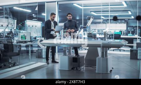 Young Engineer Works with Colleague Robotics Scientist with Laptop Computer. They Collaborate on a Robot Arm Development in an Industrial Startup. Analyzing and Evaluating Technology Systems. Stock Photo