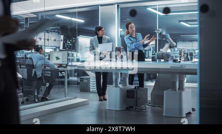 Two Female Working in High Tech Facility. Robotics Engineer and Machinery Operator Collaborate on a Robot Arm Project, Using a Laptop Computer to Operate the Robot Arm to Move Around a Microchip. Stock Photo