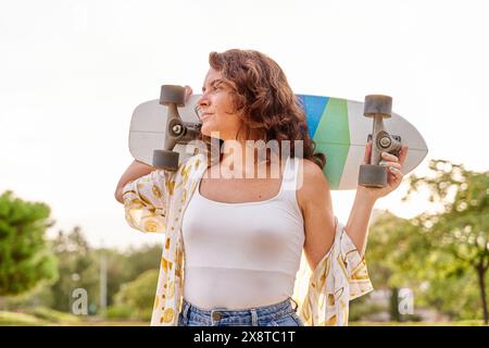 Happy skater girl walking towards the camera, sunbeams shining at lense. Young woman skating on cruiser, holding longboard on shoulders. Stock Photo