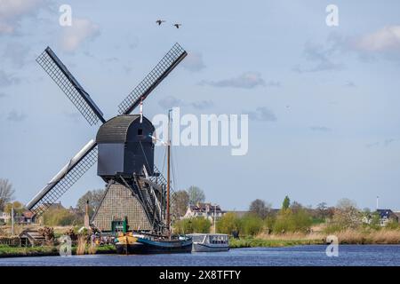 Windmill on the river bank with boats and grasses in the foreground, birds flying over the blue sky with scattered clouds, historic windmills on a Stock Photo