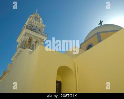 A church with a bell tower and a cross on the dome shining in yellow colour, yellow house with white tower against blue sky, Santorini, Greece Stock Photo