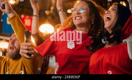 Portrait of an Anxious Multiethnic Female in Red Jersey Holding a Smartphone, Nervous About the Sports Bet on Her Favorite Soccer Team. Happy Victorious Emotions When Football Team Scores a Goal. Stock Photo