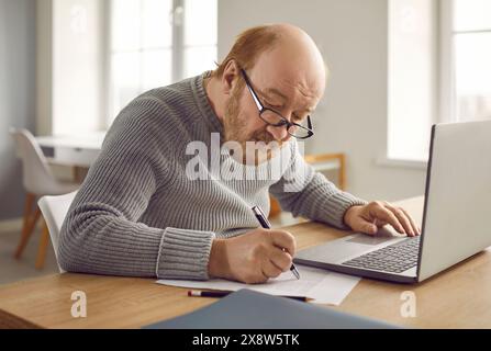 Focused senior man making notes on paper and working with laptop Stock Photo