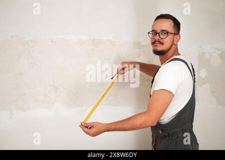 Young man with measuring tape on white background. Copy space. Smiling bearded construction worker in overalls, professional image with tape measure Stock Photo