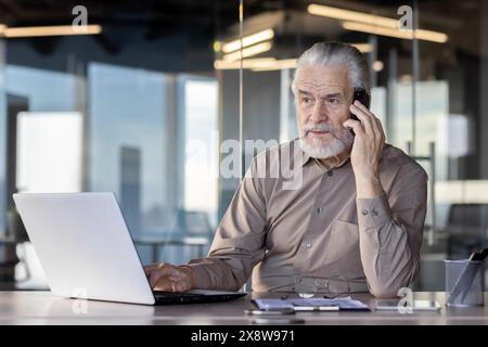 A senior professional is multitasking, talking on the phone and using a laptop in a contemporary office environment Stock Photo