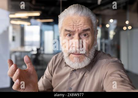 An elderly man with gray hair and beard looks surprised, gesturing towards the camera in an indoor office setting Stock Photo