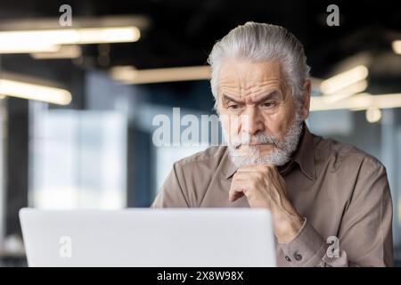 A senior man is deeply focused while using a laptop in a modern office, showing concentration and professionalism Stock Photo