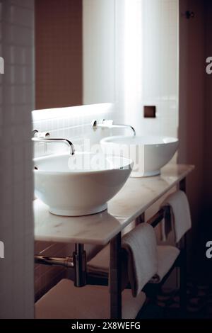 Close-up vertical shot of a modern bathroom with two white ceramic vessel sinks and chrome faucets on a marble countertop. The background features whi Stock Photo