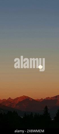 Full moon rising over a mountain range during twilight, with a gradient sky transitioning from blue to orange Stock Photo