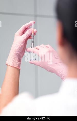 Woman cosmetologist checking for air bubbles in syringe. Back view of anonymous doctor in pink gloves holding injection preparing for beauty treatment in clinic Stock Photo